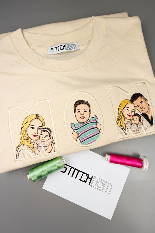 Tricou crem din bumbac organic, împăturit, cu poze brodate personalizate care formează textul „MOM”, cu portrete de familie desenate în interiorul literelor, alături de două conuri de ață verde și roz și un card alb cu logo-ul „Stitchdom”, pe un fundal gri.
