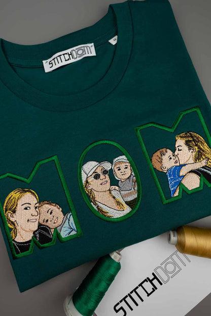 Tricou verde petrol închis din bumbac organic, împăturit, cu portrete de familie brodate în interiorul literelor „MOM”, cu brandul „STITCHDOM” vizibil pe eticheta interioară de la gât și pe un card de prezentare.