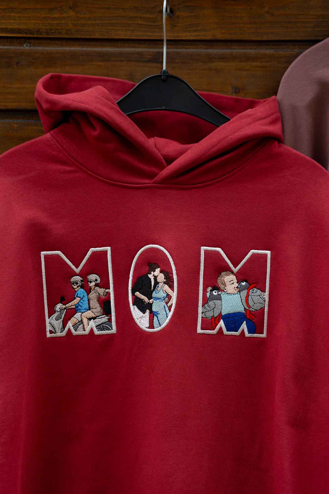 Hanorac roșu cu glugă, pe umeraș, cu textul „MOM” brodat, fiecare literă conținând câte o ilustrație brodată, pe fundal din lemn.
