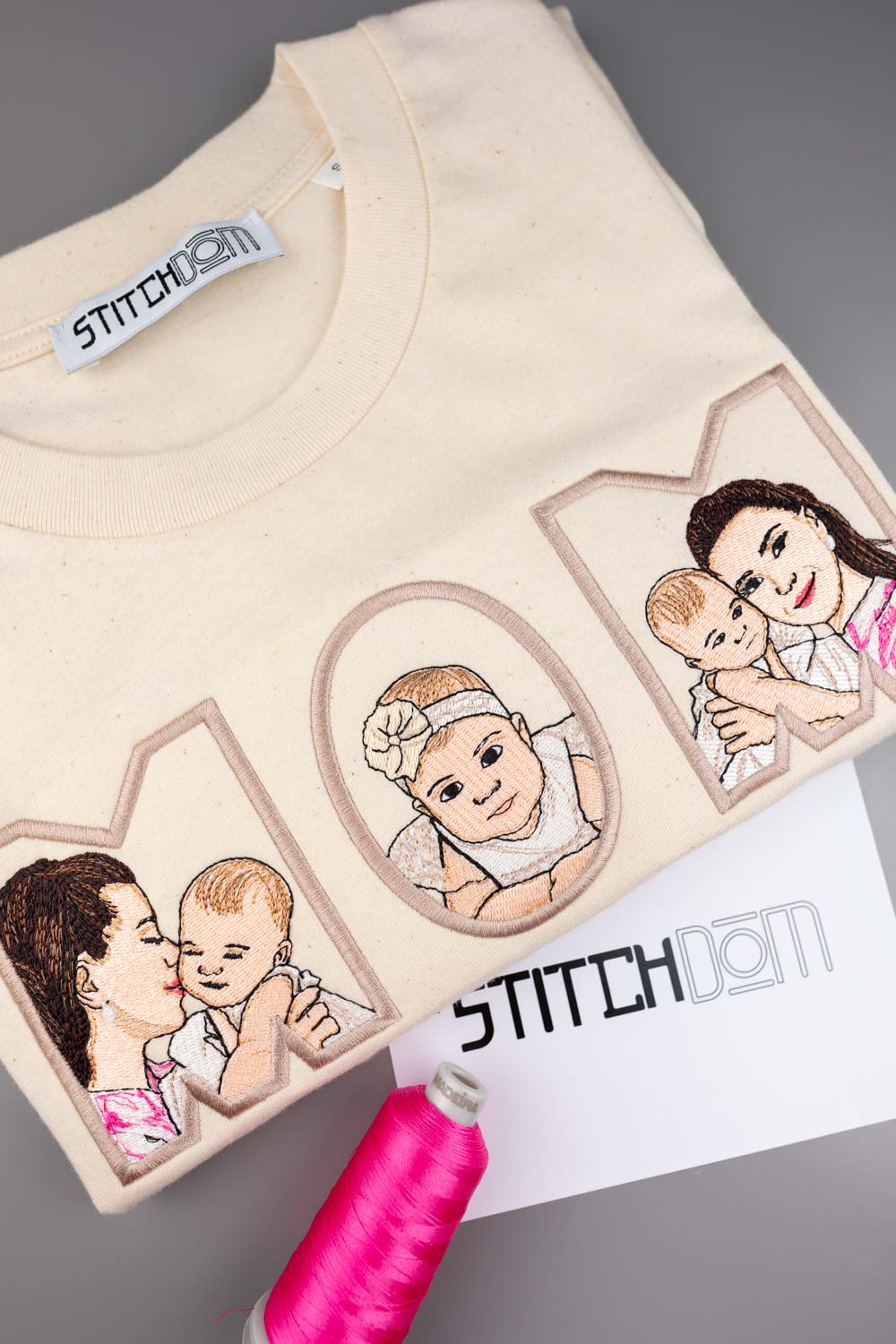 Tricou crem din bumbac organic, împăturit, cu textul „MOM” brodat personalizat, având în interior portrete ilustrate brodate ale unei mame și ale bebelușului, alături de un con de ață roz și un card cu brandul „Stitchdom”, pe un fundal gri.