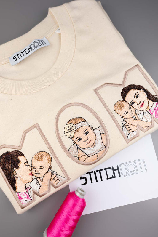 Tricou crem din bumbac organic, împăturit, cu textul „MOM” brodat personalizat, având în interior portrete ilustrate brodate ale unei mame și ale bebelușului, alături de un con de ață roz și un card cu brandul „Stitchdom”, pe un fundal gri.