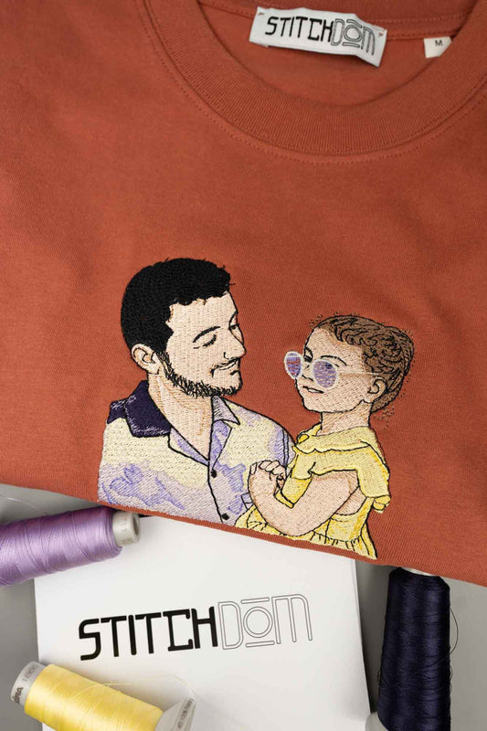 Tricou din bumbac organic cu model brodat reprezentând un bărbat și o femeie, pe un fundal asortat, cu brandul „Stitchdom" vizibil.