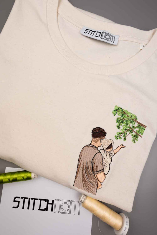 Tricou crem din bumbac organic, împăturit, de la StitchDom, cu o poză brodată cu un bărbat care ține un bebeluș lângă o ramură verde de copac, pe un fundal gri, alături de accesorii de cusut, inclusiv un con de ață.