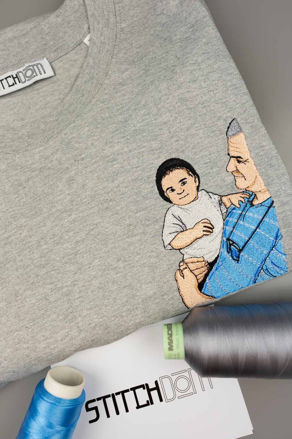 Tricou gri deschis din bumbac organic, împăturit, cu o poză brodată colorată în stil desen, cu un bărbat care ține un copil în brațe, alături de conuri de ață și o etichetă de brand „Stitchdom”, pe un fundal gri.