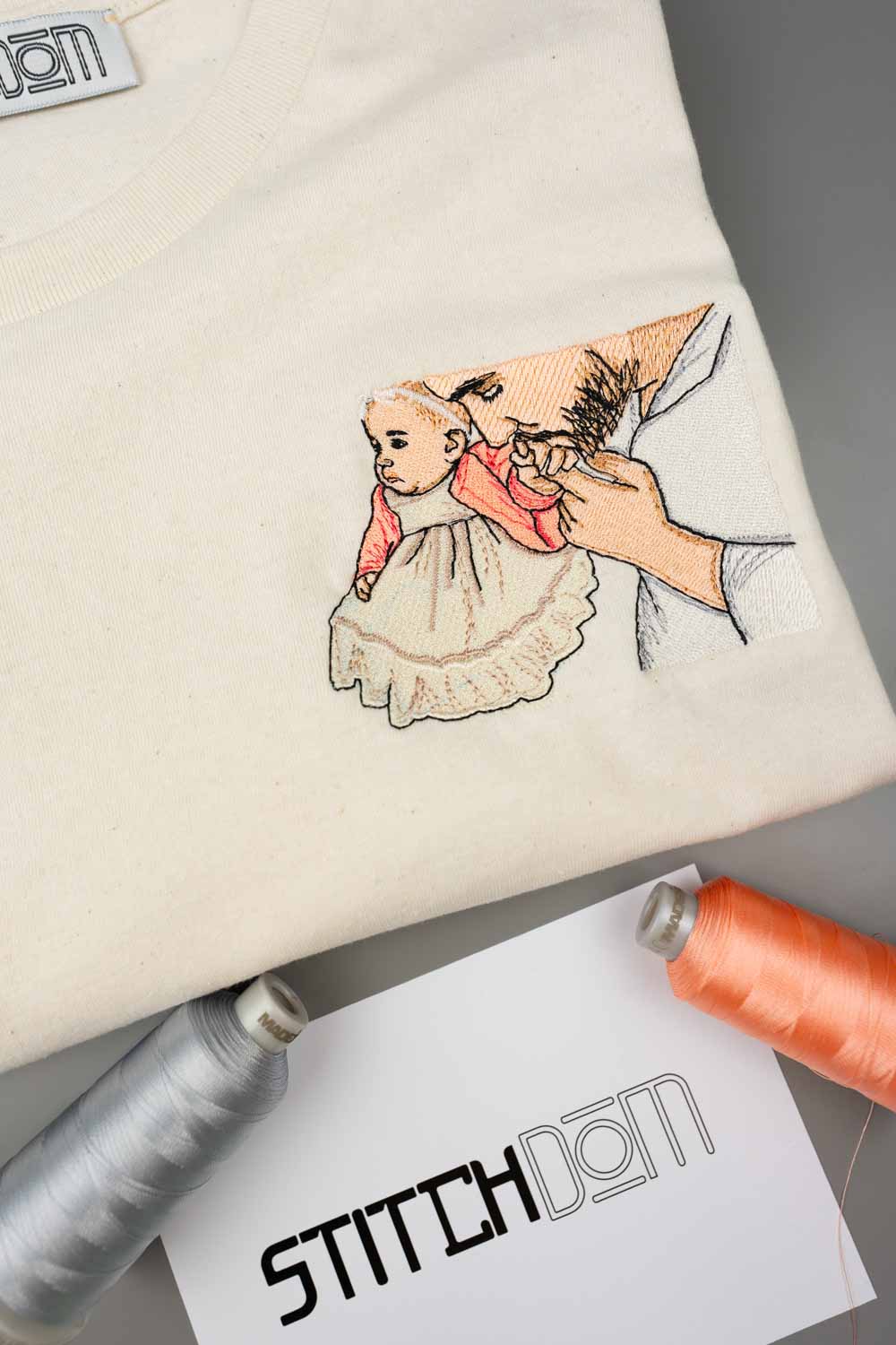 Tricou crem din bumbac organic, împăturit, cu o poză brodată detaliată cu un tata care își ține bebelușul în brațe, alături de conuri de ață și un card de brand „Stitchdom”.