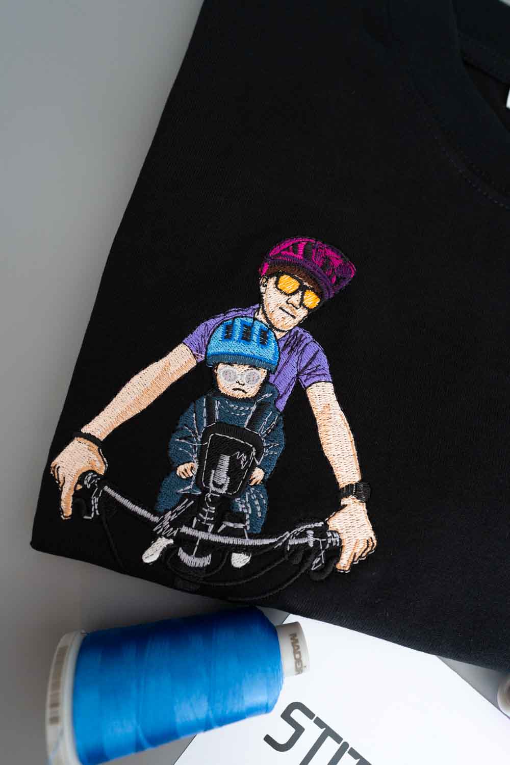Tricou negru din bumbac organic, așezat flat lay, cu o poză brodată colorată cu un tata si copilul lui care merg pe bicicletă, alături de un con de ață albastră.