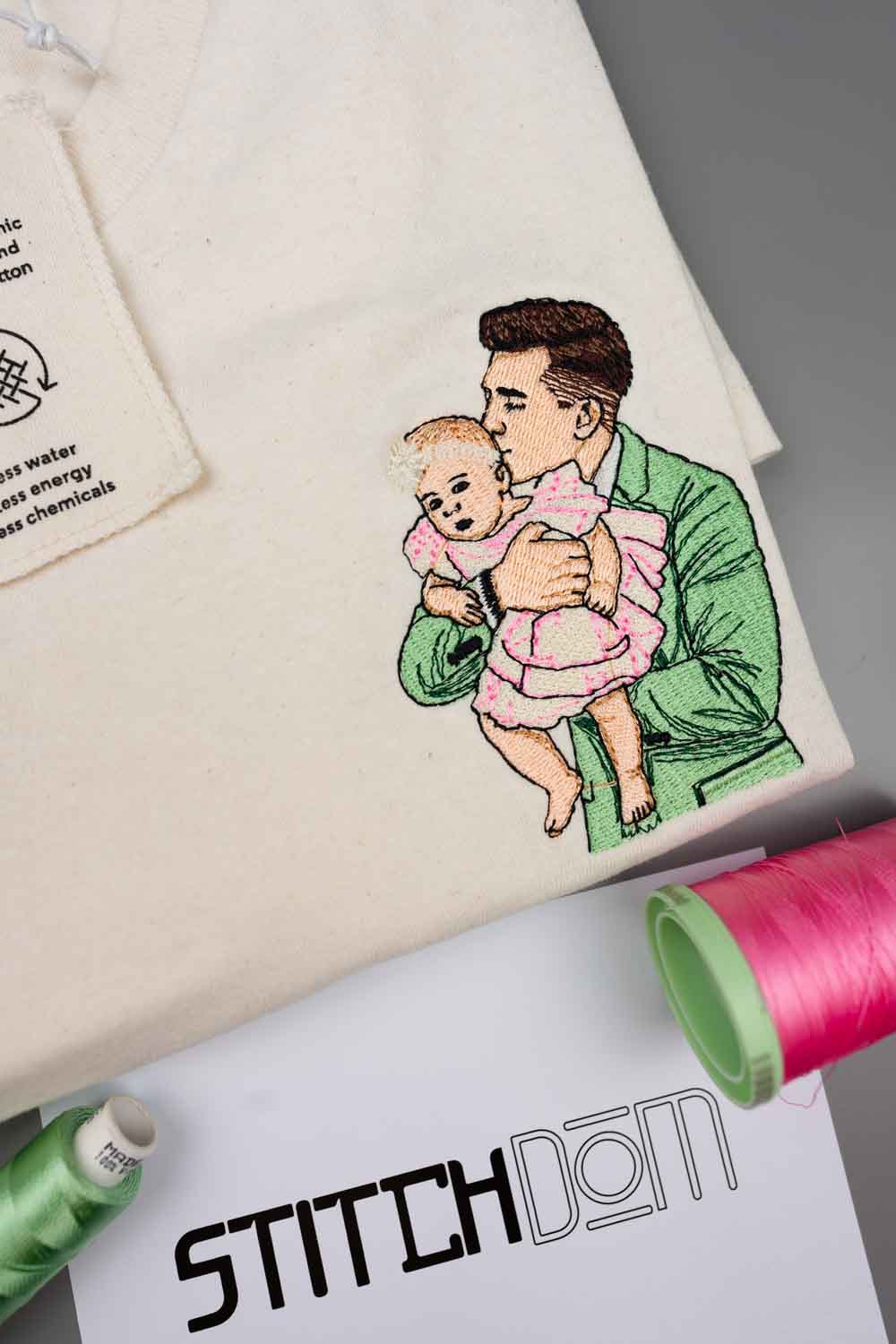 Tricou crem, împăturit, cu o poză brodată, colorată, cu un bărbat îmbrăcat într-un sacou verde care ține în brațe un bebeluș îmbrăcat într-o rochiță roz, pe o suprafață gri, alături de conuri de ață verde și roz și o foaie albă cu brandul „STITCHDOM”.