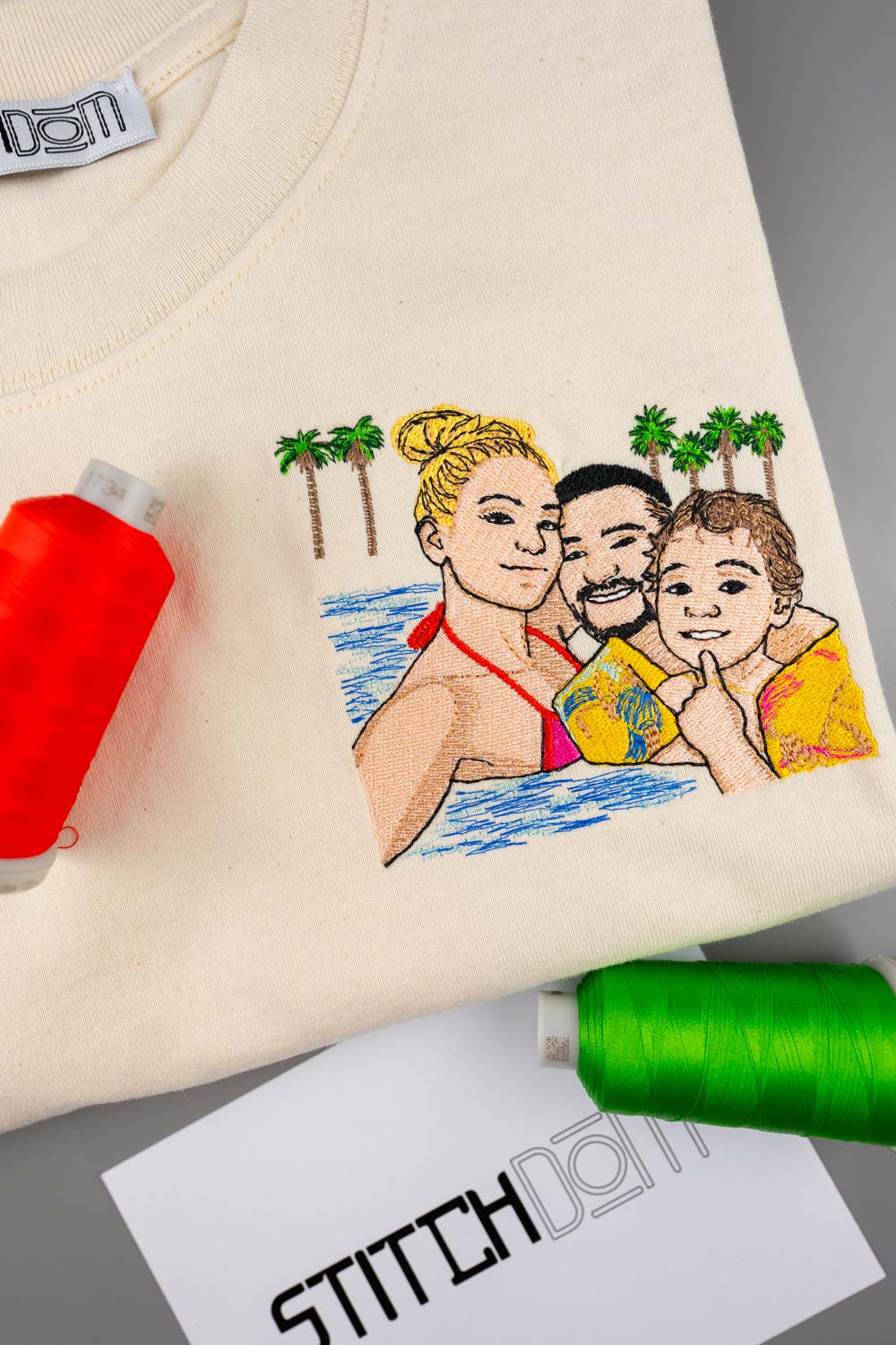 Tricou crem din bumbac organic, cu un portret de familie colorat brodat, având palmieri în fundal, alături de conuri de ață roșie și verde și un card cu brandul „Stitchdom”, pe o suprafață gri.