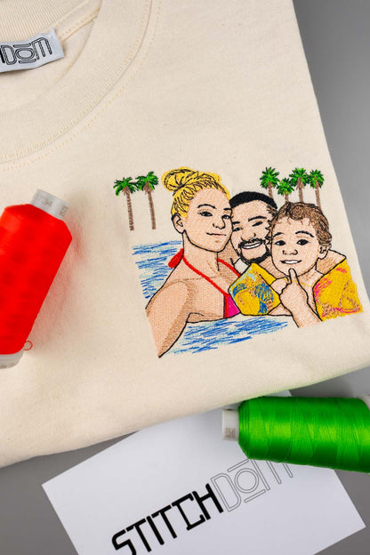 Tricou crem din bumbac organic, cu un portret de familie colorat brodat, având palmieri în fundal, alături de conuri de ață roșie și verde și un card cu brandul „Stitchdom”, pe o suprafață gri.