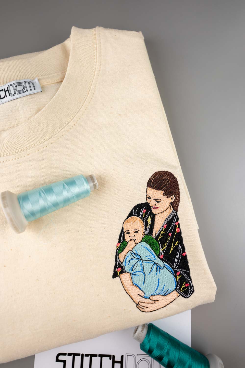 Tricou crem din bumbac organic, împăturit, cu o ilustrație colorată brodată reprezentând o mamă care își ține bebelușul în brațe, alături de conuri de ață bleu deschis și verde, pe un fundal gri neutru.