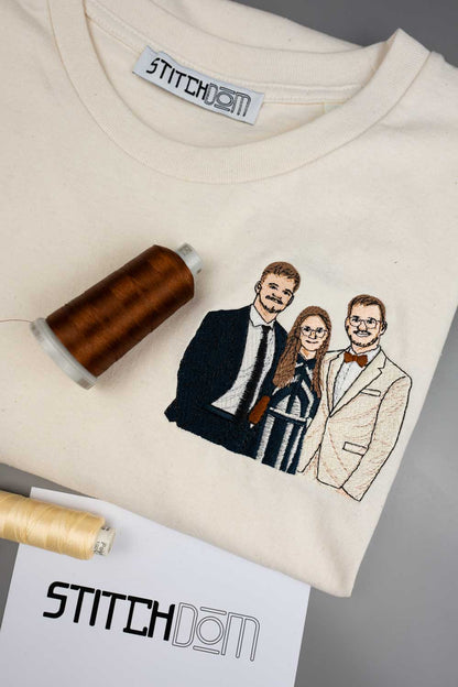 Tricou alb din bumbac organic, cu guler rotund, având o ilustrație personalizată brodată cu trei personae pe partea din față, alături de două conuri de ață, unul maro și unul bej, și un card cu brandul „Stitchdom”, pe un fundal gri.