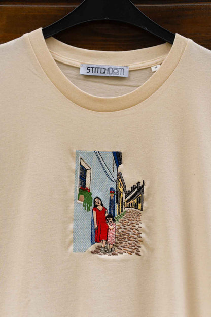 Tricou bej din bumbac cu model brodat reprezentând o femeie într-o rochie roșie pe o stradă.