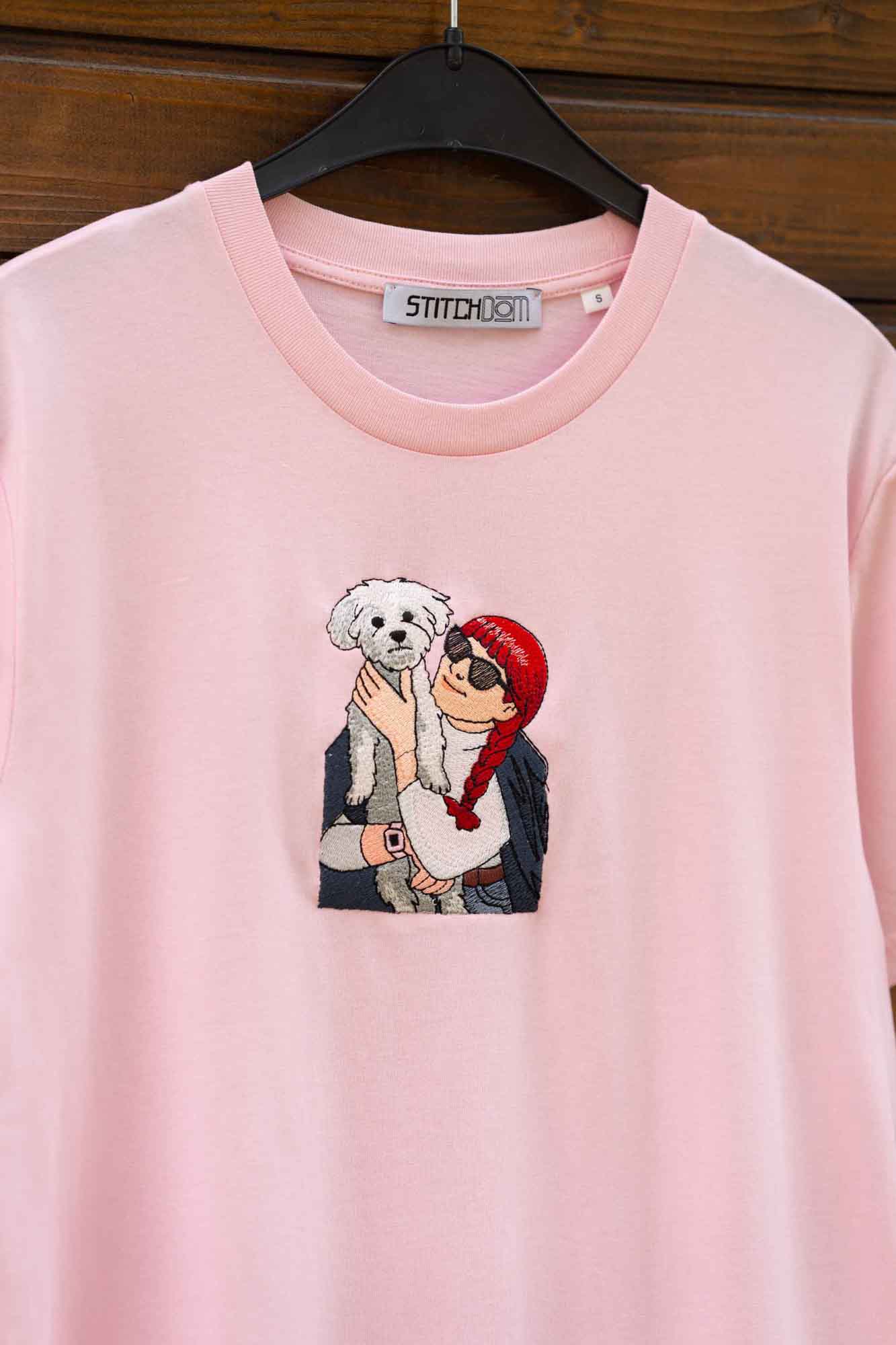 Tricou roz cu model brodat reprezentând o persoană care ține un câine, pe o suprafață din lemn.