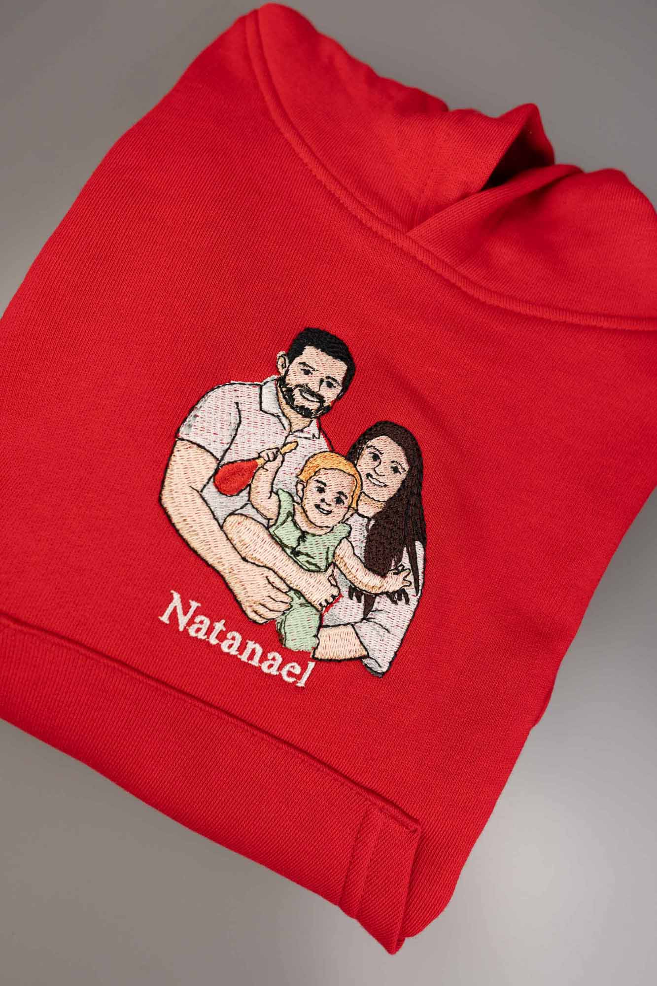 Hanorac roșu din bumbac organic, împăturit, pe un fundal gri neutru, cu o poză de familie brodată personalizată și numele „Natanael”.