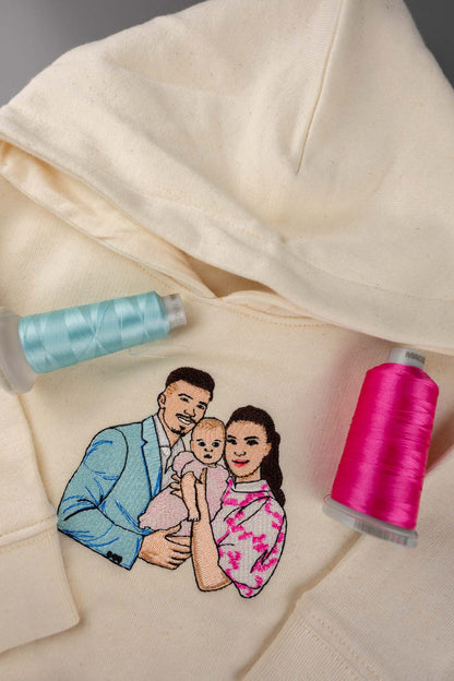 Hanorac crem din bumbac organic, prezentat în prim-plan, cu o ilustrație personalizată brodată reprezentând o familie formată din trei persoane, alături de două conuri de ață, unul bleu deschis și unul roz aprins, așezate de-o parte și de alta a broderiei.