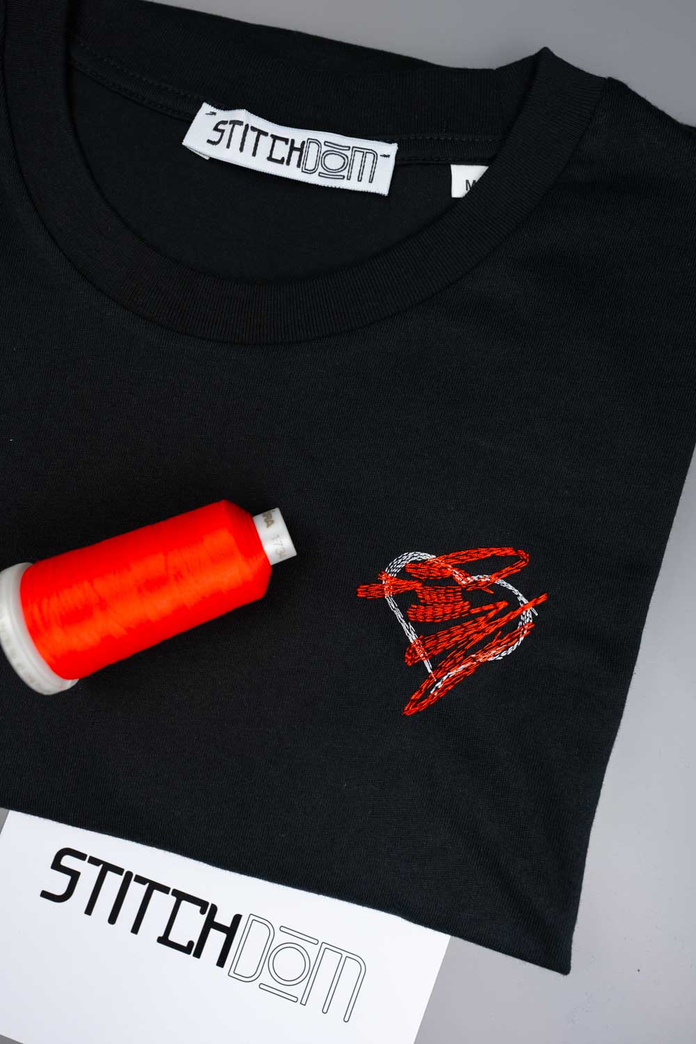 Tricou negru din bumbac organic de la Stitchdom, cu un design brodat în formă de inimă, în nuanțe de roșu și alb, pe piept, alături de un con de ață portocaliu aprins și un card de hârtie cu brandul, pe un fundal gri.