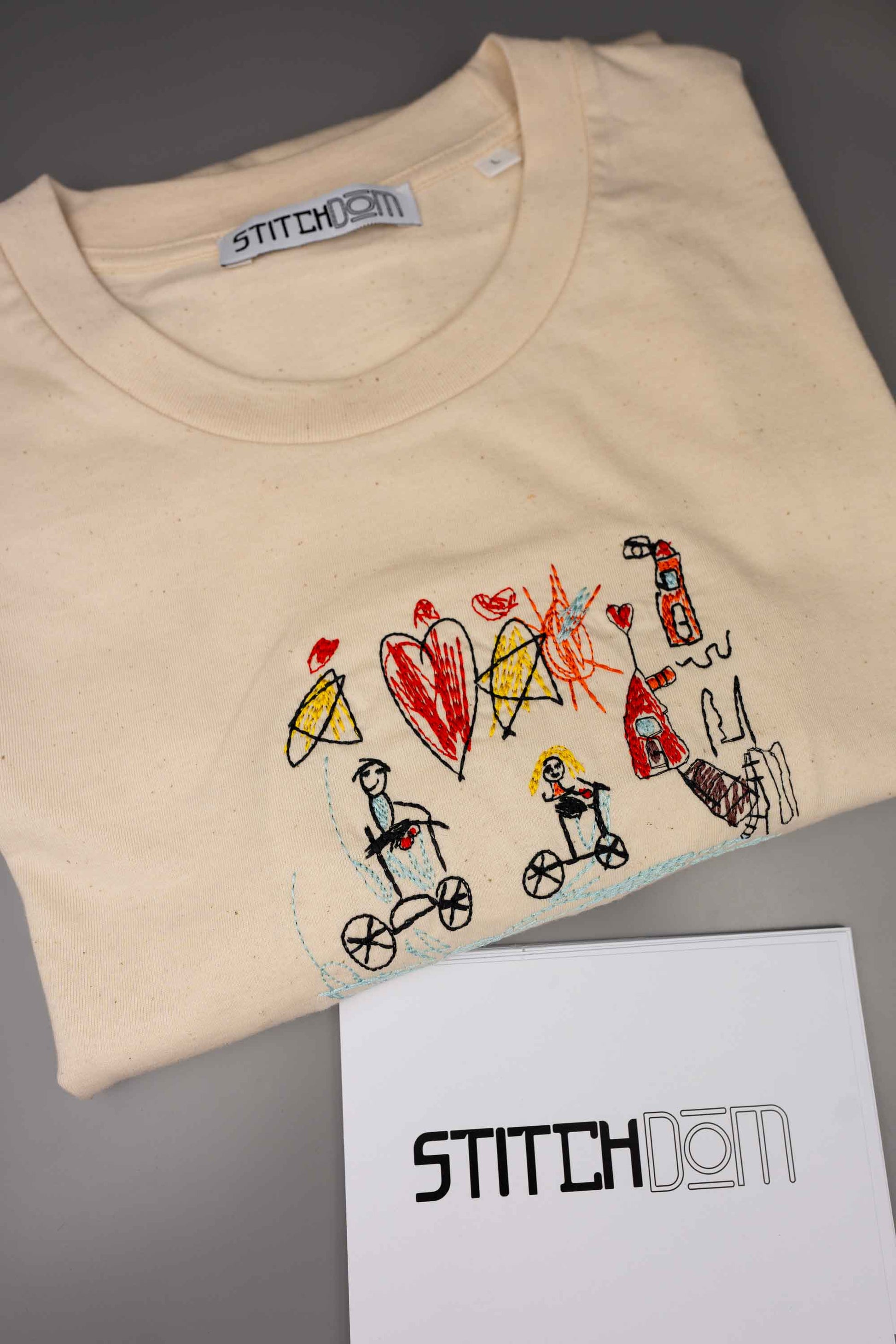 Tricou crem din bumbac organic, împăturit, cu o poză brodată colorată cu desen de copii, cu personaje pe biciclete, inimioare și o casă, pe o suprafață albă, cu textul „STITCHDOM” vizibil.
