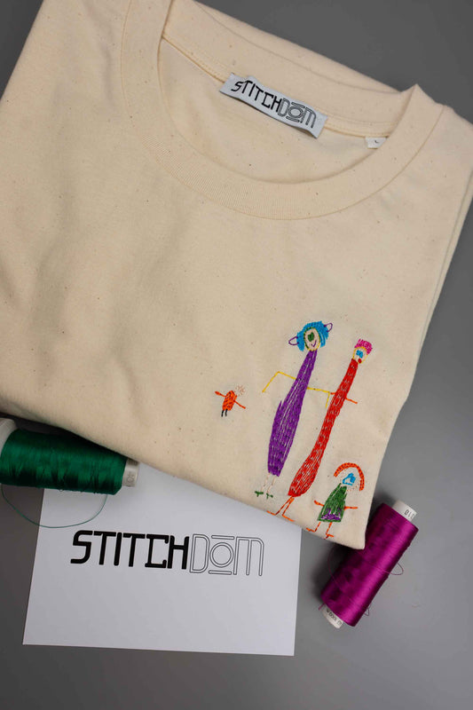 Tricou crem din bumbac organic, împăturit, cu poze brodate colorate, alături de conuri de ață verde, mov și alb, precum și un card „Stitchdom”, pe o suprafață gri.