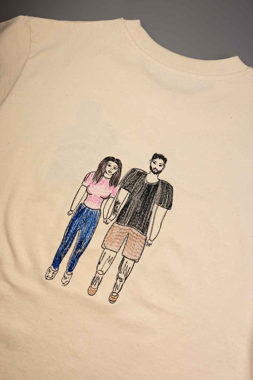 Tricou crem din bumbac organic, cu o poză brodată în stil desen de mână, reprezentând un cuplu pe partea din față, pe un fundal gri neutru.