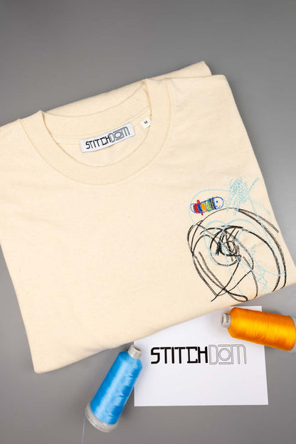 Tricou crem din bumbac organic, împăturit, cu o poză brodată colorată reprezentând desenul copilului lor, însoțit de conuri de ață și un card cu brandul „STITCHDOM”, pe un fundal gri.
