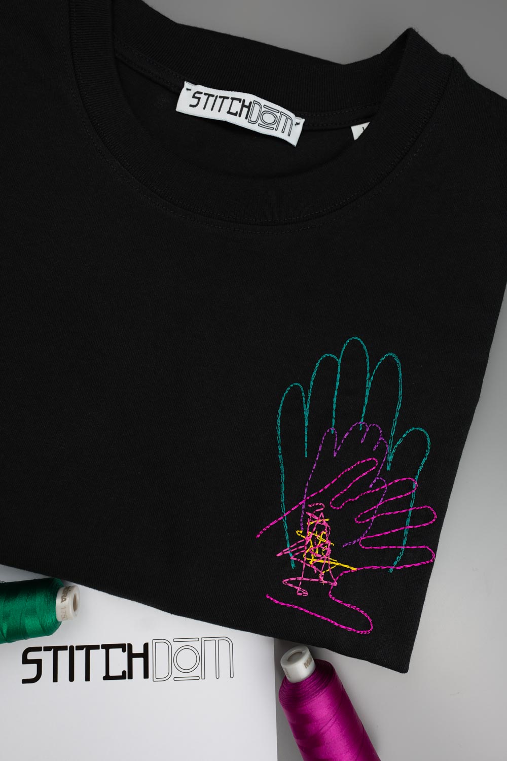 Tricou negru din bumbac organic, cu guler rotund, așezat pe o suprafață gri deschis, cu o poză brodată colorată reprezentând o mână realizată din bucle de ață în nuanțe de verde petrol, roz și galben, alături de conuri de ață în colțurile de jos.