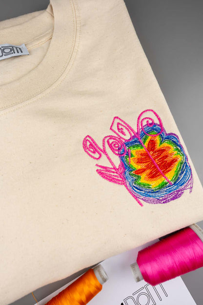 Tricou crem din bumbac organic, împăturit, cu un design colorat brodat pe piept, format din conturul unei mâini roz care încadrează un model vibrant de frunze multicolore în nuanțe de verde, galben, portocaliu și roșu.