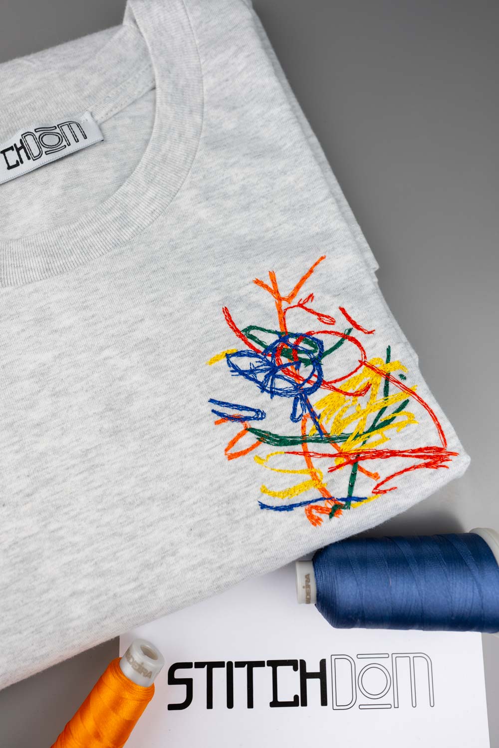 Tricou gri deschis melanj din bumbac organic, împăturit, cu un design colorat brodat tip mâzgăleală pe piept, alături de conuri de ață și un card cu logo-ul „Stitchdom”, pe un fundal gri.