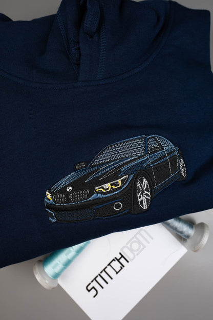 Hanorac bleumarin din bumbac organic, împăturit, cu o poză brodată cu o mașină sport neagră pe piept, alături de conuri de ață și un card alb cu textul „STITCHDOM”, pe o suprafață gri.