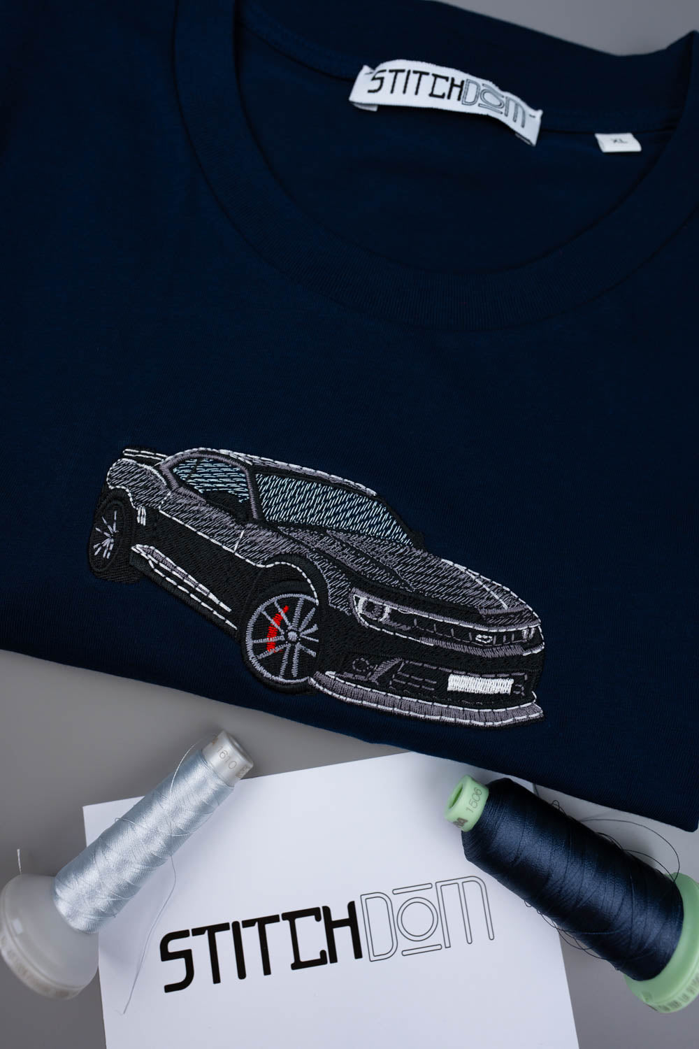 Tricou bleumarin din bumbac organic, cu o poză brodată alb-negru cu un Camaro pe piept, alături de conuri de ață și o etichetă de brand „Stitchdom”, pe o suprafață gri.