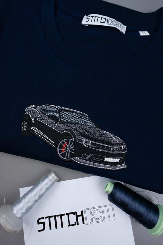 Tricou bleumarin din bumbac organic, cu o poză brodată alb-negru cu un Camaro pe piept, alături de conuri de ață și o etichetă de brand „Stitchdom”, pe o suprafață gri.