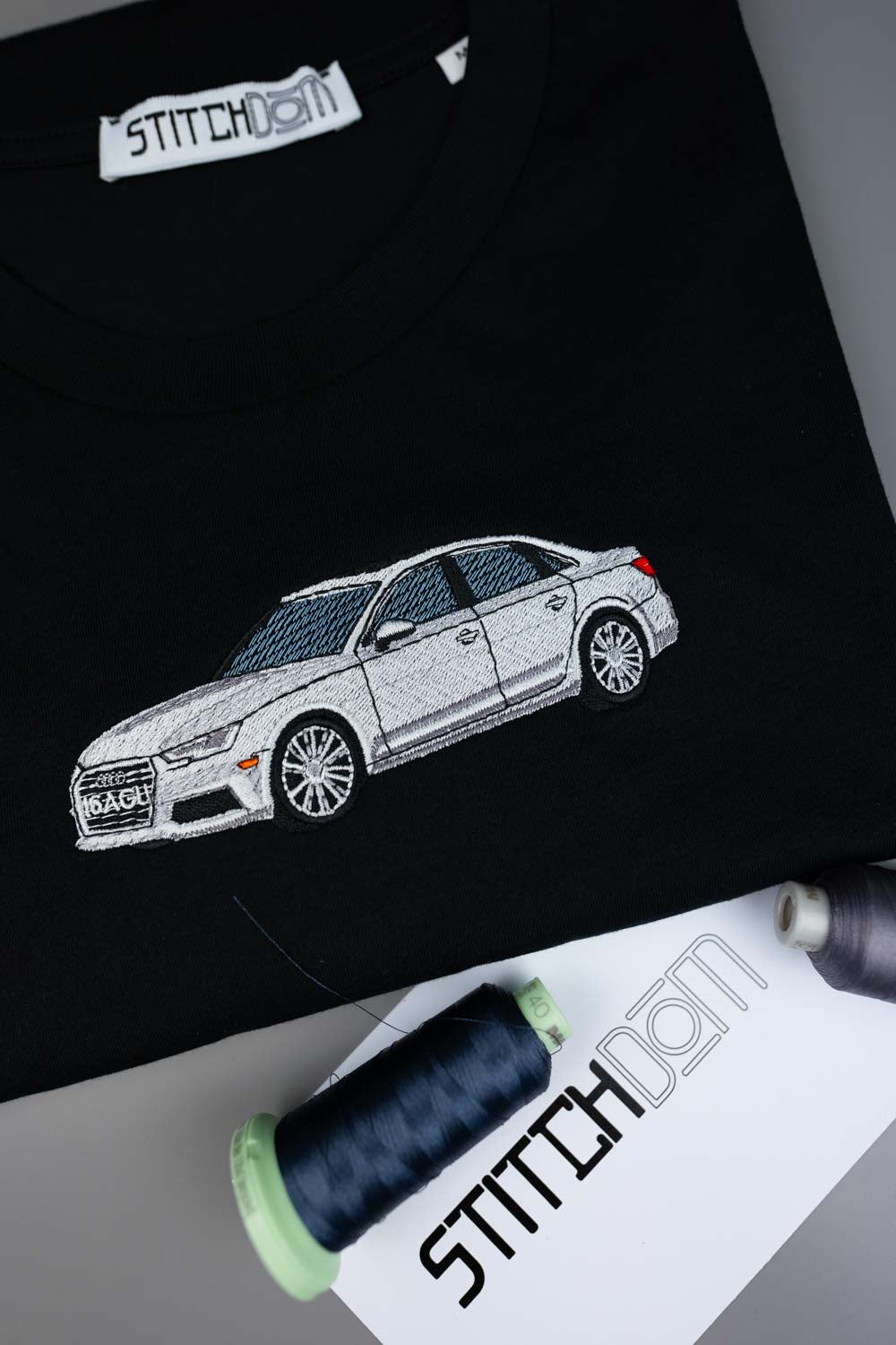 Tricou negru din bumbac organic, cu o poză brodată alb-gri în stil line art, reprezentând un sedan AUDI pe piept, aranjat flat lay, alături de conuri de ață și o etichetă albă de hârtie cu brandul „STITCHDOM”.