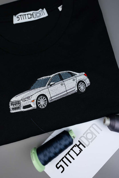 Tricou negru din bumbac organic, cu o poză brodată alb-gri în stil line art, reprezentând un sedan AUDI pe piept, aranjat flat lay, alături de conuri de ață și o etichetă albă de hârtie cu brandul „STITCHDOM”.