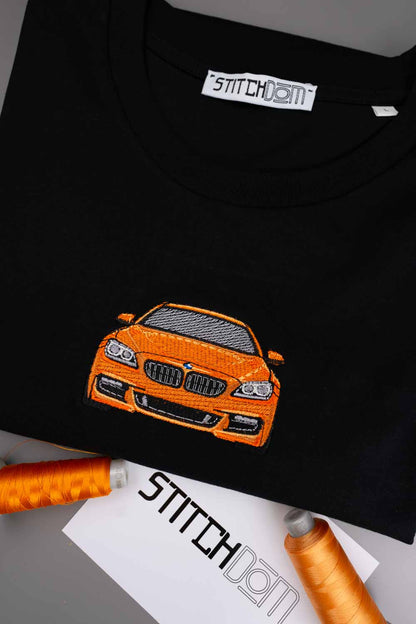 Tricou negru din bumbac organic, cu guler rotund, având o mașină sport portocalie brodată pe piept, alături de conuri de ață portocalie și o etichetă de brand „Stitchdom”.