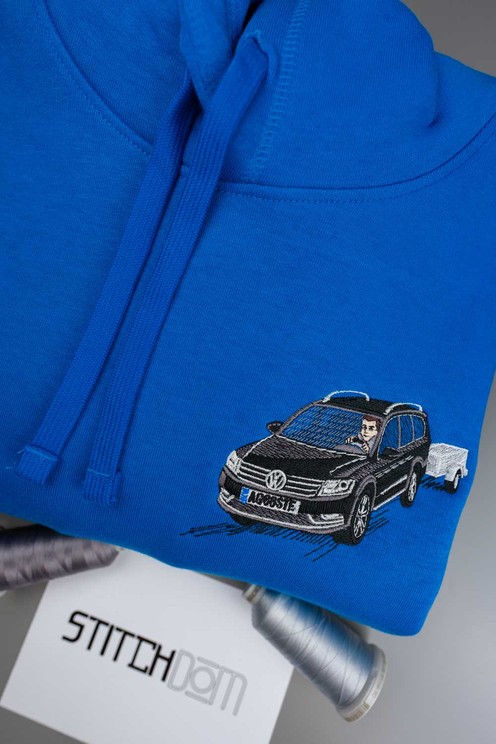 Hanorac albastru aprins din bumbac organic, împăturit, cu o ilustrație neagră brodată reprezentând un Volkswagen combi care tractează o remorcă, alături de conuri de ață și un card cu brandul „Stitchdom”.