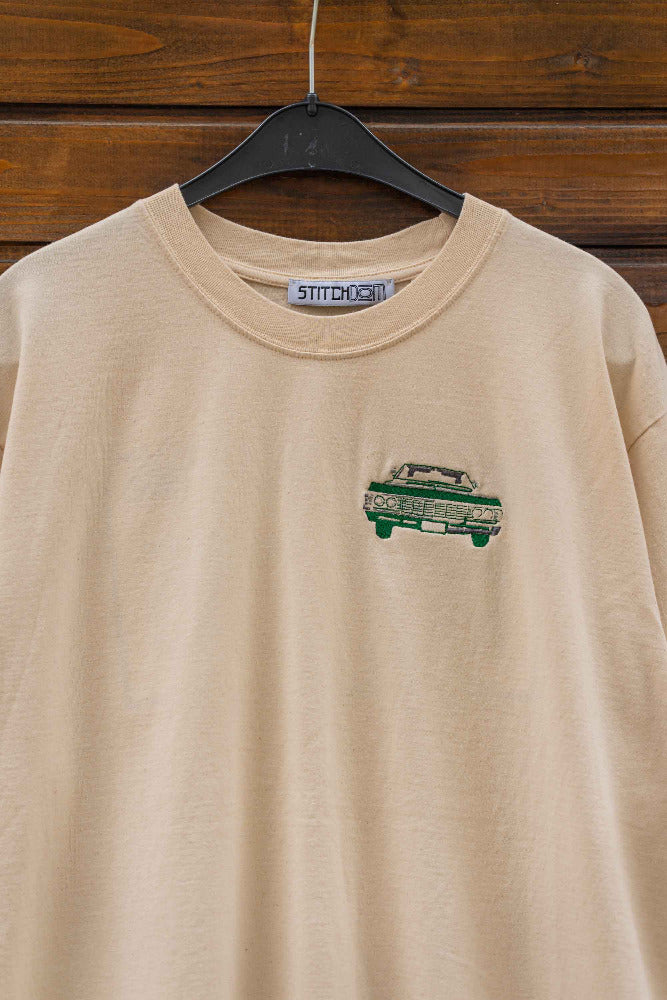 Tricou bej din bumbac organic, pe umeraș, pe un fundal din lemn, cu o masina brodată verde.