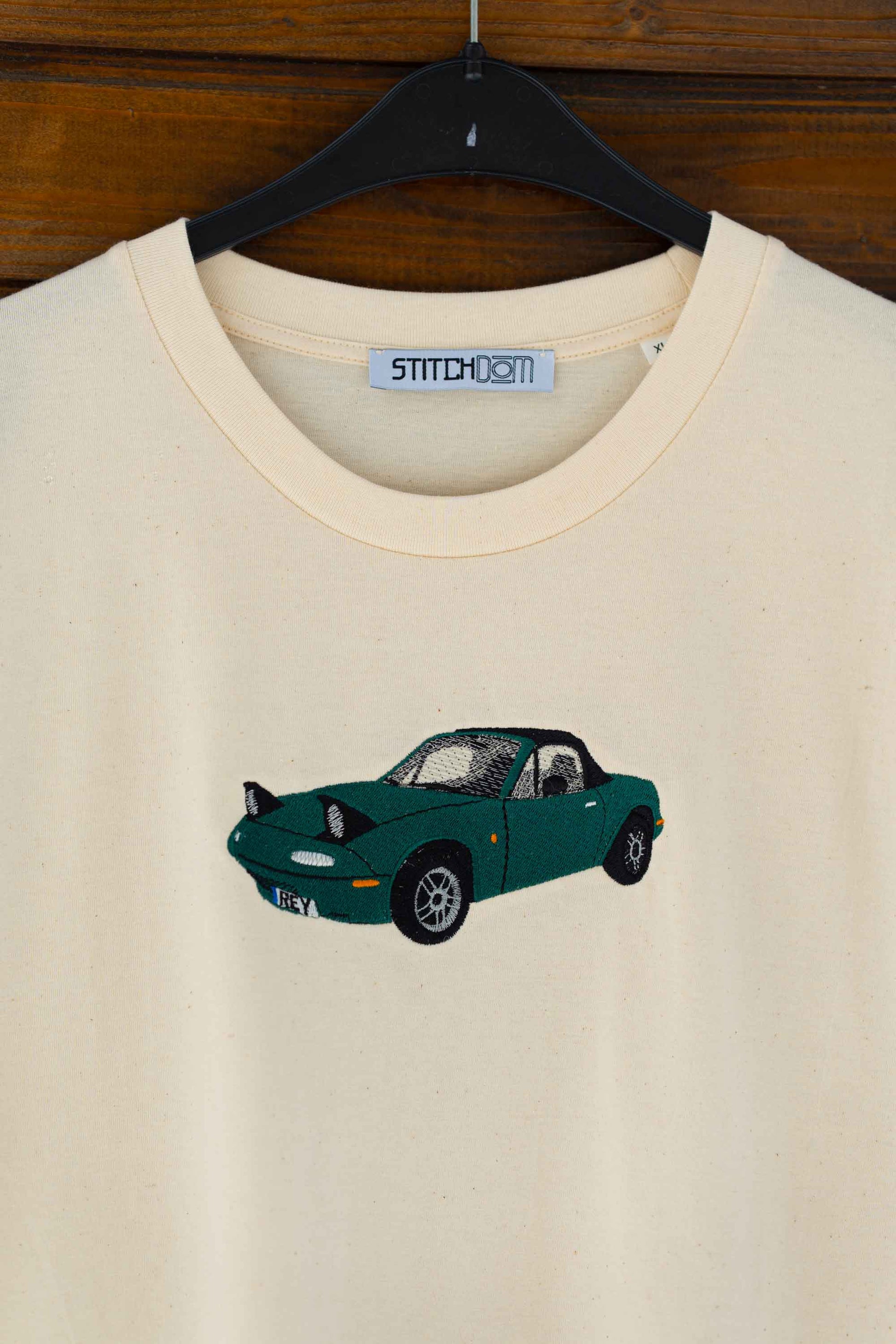 Tricou bej din bumbac organic, pe umeraș, pe un fundal din lemn, cu o poză brodată cu o mașină verde.