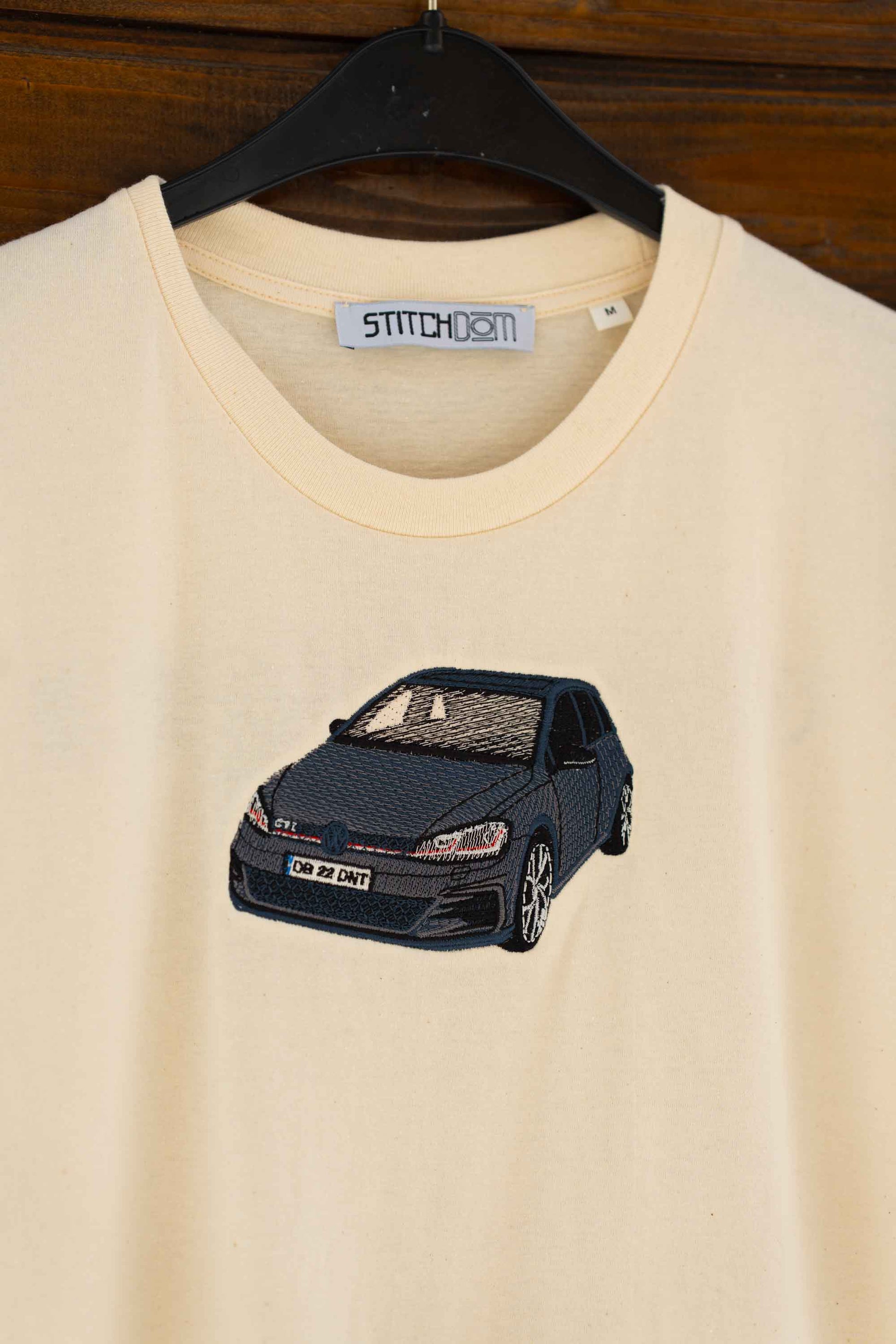 Tricou bej din bumbac organic, pe un fundal din lemn, cu o poză brodată cu o mașină neagră, un Volkswagen Golf.