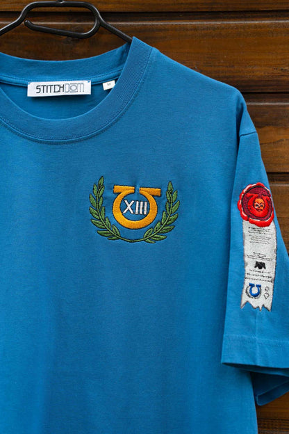 Tricou albastru aprins din bumbac organic, cu mânecă scurtă, agățat pe un umeraș negru, pe un fundal din lemn închis, cu o emblemă brodată galben-verde insiprata din Warhammer, pe partea stângă a pieptului, având cifra romană „XIII”, și un patch alb brodat pe mânecă, cu detalii roșii și albastre.