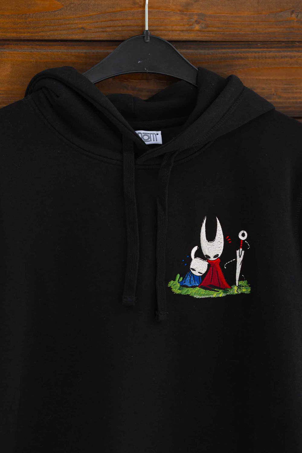 Hanorac negru din bumbac organic, agățat pe un umeraș negru, pe un fundal din lemn închis, cu un mic design brodat pe partea stângă a pieptului, reprezentând o figură albă cu glugă și robă roșie și o figură albastră pe o porțiune de iarbă verde, inspirat din jocul Silksong.