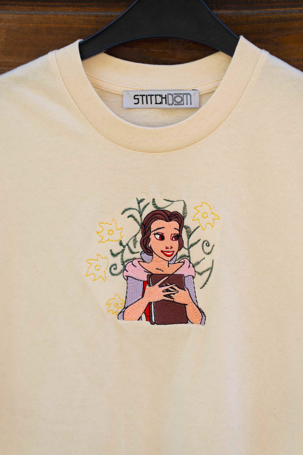 Tricou crem din bumbac organic, agățat pe un umeraș negru, pe un fundal din lemn închis, cu o poză brodată reprezentând-o pe Belle din „Frumoasa și Bestia” în centrul pieptului, înconjurată de flori galbene și vrejuri verzi brodate.