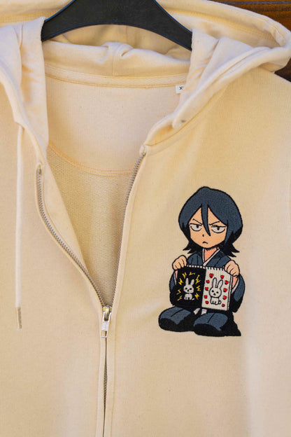Hanorac crem cu fermoar din bumbac organic, agățat pe un umeraș negru, cu un mic personaj anime goth brodat pe partea stângă a pieptului, ținând un jurnal în mână.