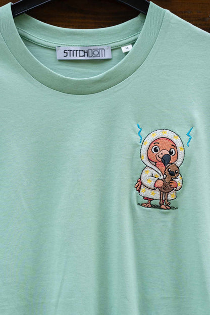 Tricou verde mentă din bumbac organic, cu guler rotund, de la Stitchdom, agățat pe un umeraș negru, cu o poză brodată reprezentând un personaj cartoon cu o pasăre dodo pe partea stângă a pieptului.