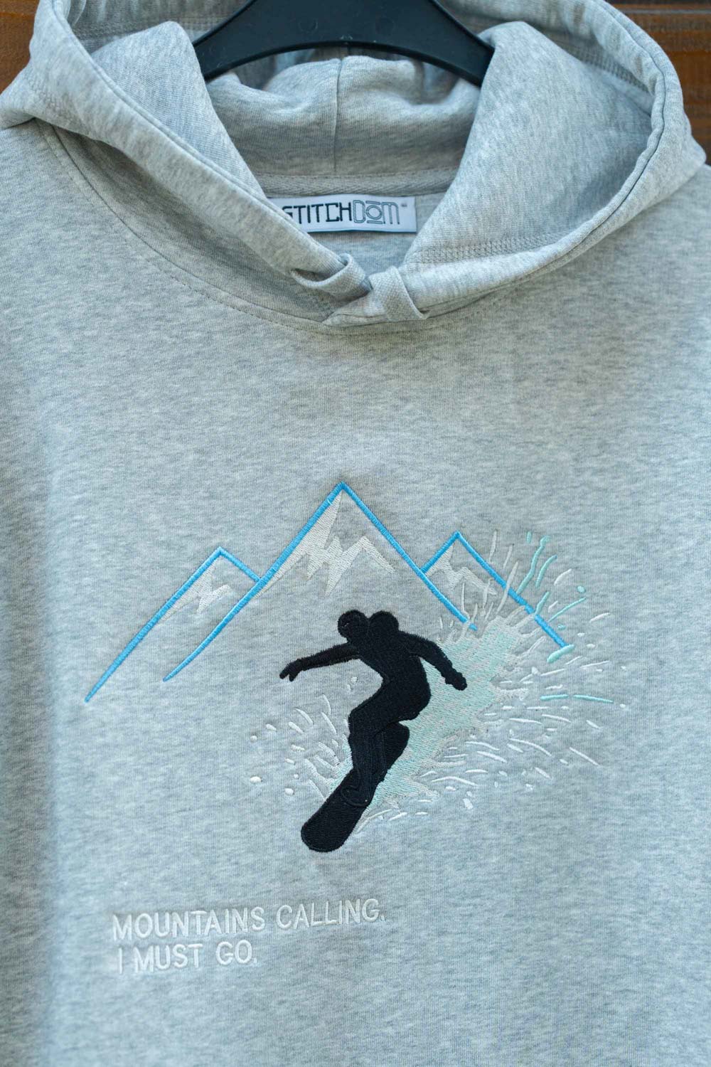 Hanorac gri deschis melanj din bumbac organic, agățat pe un umeraș negru, cu o poză brodată reprezentând un snowboarder negru, înconjurat de linii albastre și albe ce formează munți, și textul „MOUNTAINS CALLING, I MUST GO.” brodat.