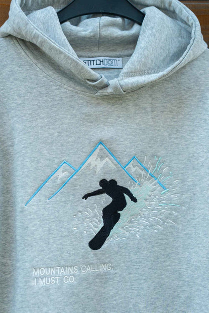 Hanorac gri deschis melanj din bumbac organic, agățat pe un umeraș negru, cu o poză brodată reprezentând un snowboarder negru, înconjurat de linii albastre și albe ce formează munți, și textul „MOUNTAINS CALLING, I MUST GO.” brodat.