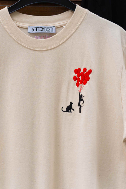 Tricou crem din bumbac organic, cu guler rotund, agățat pe un umeraș negru, cu un design brodat pe partea stângă a pieptului, reprezentând o pisică neagră și o siluetă mică ce ține baloane roșii, cu eticheta de brand „STITCHDOM” vizibilă în interiorul gulerului.