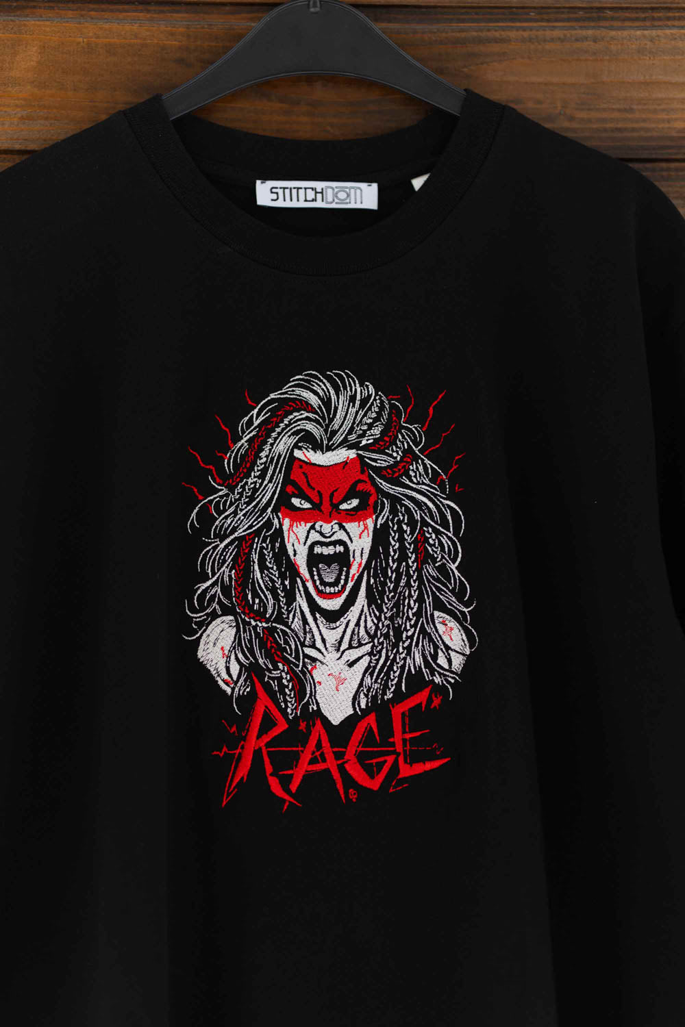 Tricou negru din bumbac organic, agățat pe un umeraș, cu o poză brodată reprezentând chipul unei femei și textul „RAGE” brodat pe piept.