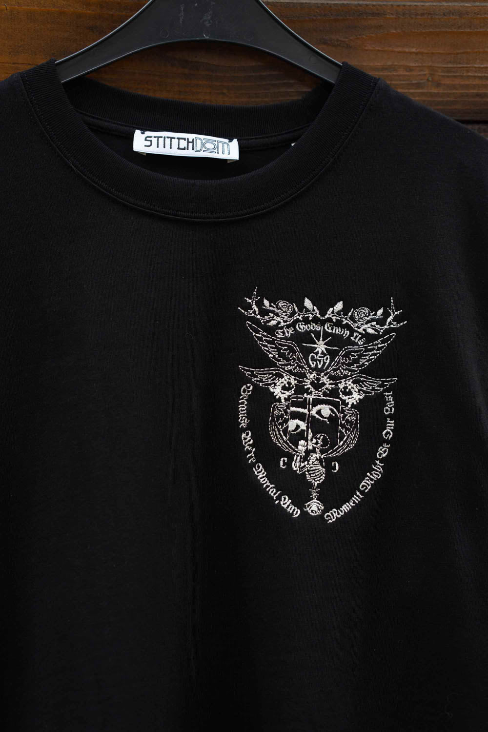 Tricou negru din bumbac organic, agățat pe un umeraș, cu un design grafic alb brodat pe piept.