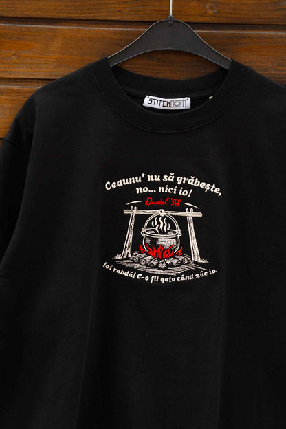 Tricou negru din bumbac organic, agățat pe un umeraș, pe un fundal din lemn, cu un design grafic brodat și text brodat pe piept.