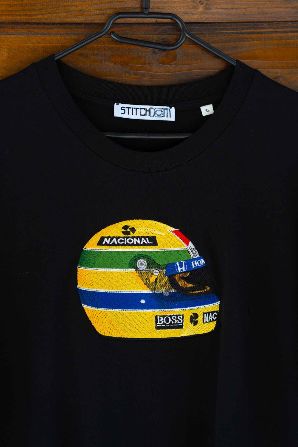 Tricou negru din bumbac organic, cu guler rotund, agățat pe un umeraș negru, pe un fundal din lemn, cu un patch brodat colorat reprezentând o cască de curse în nuanțe de galben, verde, albastru și alb, cu textele „NACIONAL” și „BOSS” vizibile pe patch.