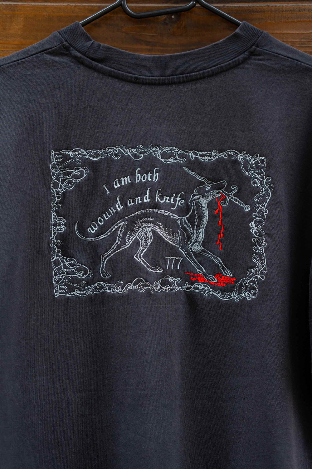Tricou negru din bumbac organic, pe un umeraș negru, pe un fundal din lemn închis, cu o poză brodată reprezentând un lup într-o ramă decorativă, alături de textele „I am both wound and knife” și „777” brodate cu alb și detalii roșii în formă de picături de sânge.
